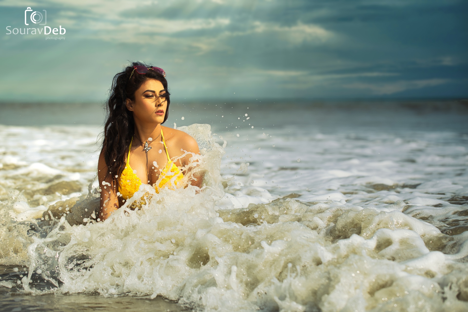 This photograph of model Soumi Das, captured by the talented photographer Sourav Deb for his 2020-21 Swimsuit Calendar, is a captivating portrait of feminine beauty amidst the ocean's embrace. The image features a woman kneeling in the shallows, her yellow bathing suit a vibrant splash against the turquoise water. Sparkling droplets of seawater cascade down her skin, capturing the playful essence of the ocean's spray.The composition draws the viewer's eye to the woman's face, partially turned towards her left. Sunlight bathes her features, creating a striking Rembrandt shadow that adds depth and dimension to the image. Above the waterline, the bikini top and a glimpse of her shoulder emerge, further emphasizing the feminine form.Sourav Deb's masterful use of high dynamic range (HDR) photography enhances the image's visual impact. The vibrant colors of the ocean and the woman's swimsuit stand in stark contrast to the deep shadows, creating a sense of drama and intrigue. The overall effect is a stunning and captivating portrait that celebrates both the beauty of the female form and the power of the natural world.
